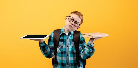 Confused kid schooler with backpack and glasses holding digital tablet and book, choosing between...