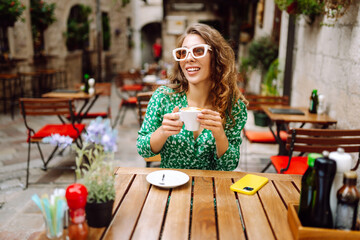 Portrait of happy woman drinking cup of coffee at wooden table on cozy veranda. Young tourist enjoying morning and hot drink outdoors. Tourism and drinks concept.