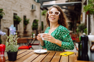 Portrait of happy woman drinking cup of coffee at wooden table on cozy veranda. Young tourist enjoying morning and hot drink outdoors. Tourism and drinks concept.