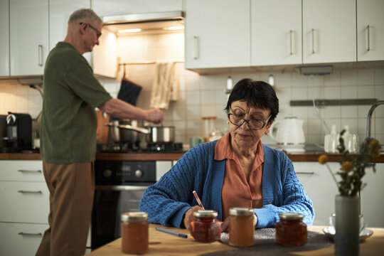 Elderly couple seen in cozy kitchen making summer preserves and writing labels for jam jars. Background features modern appliances and wooden countertops - Powered by Adobe
