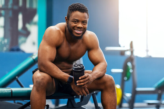 Smiling african american shirtless bodybuilder drinking protein or water while having break after workout at modern gym. Happy black guy sportsman resting after exercising, copy space