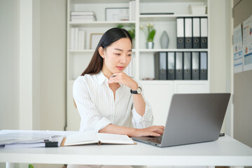 Obraz premium Focused Asian businesswoman working on a laptop in a bright office, looking engaged with her task.