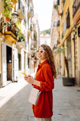 Portrait of young woman with phone walking along sunny city streets. Beautiful woman enjoying...