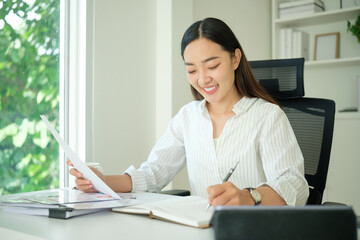 Focused businesswoman wearing glasses reviewing a document and writing notes while working at her office desk.
