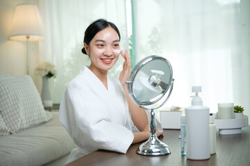 Cheerful woman applying moisturizing cream to her face while looking in the mirror during skincare...