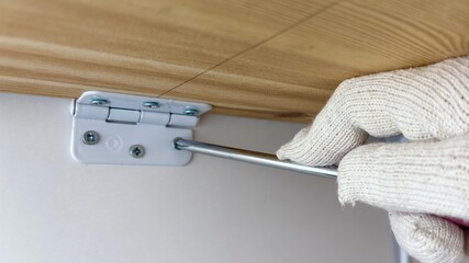 Gloved hand uses a screwdriver to install a hinge on a wooden surface