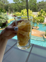 Fresh Pineapple in a Plastic Cup Held by Hand Against Pool and Palm Trees Background &ndash; Healthy Summer Snack on Vacation.Sliced pineapple in a clear plastic cup held in a woman&rsquo;s hand.
