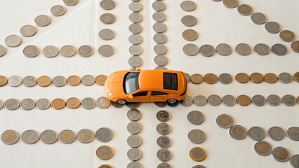 Miniature orange car is placed white surface surrounded by arranged coins forming intersecting paths, symbolizing financial decisions and planning