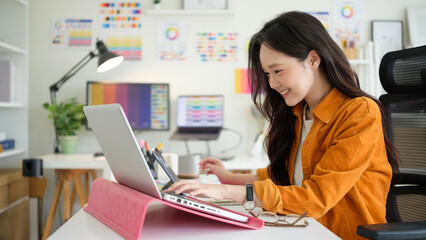 Smiling young female graphic designer working on a laptop in a creative home office.