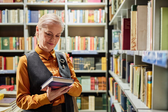 Senior Caucasian woman standing in library holding books, smiling while reading cover, surrounded by shelves filled with books, appearing engaged in choosing literature