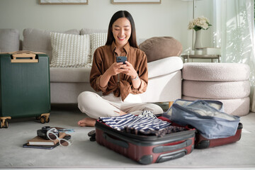 Smiling woman using a smartphone while preparing for her trip, sitting on the floor with a suitcase...