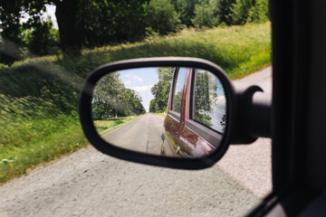 Car side mirror reflection. Rural road with trees and green field. Driving through countryside in summer. View from passenger window. Safe travel in natural scenic environment.