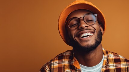 Fototapeta premium A joyful young man donned in an orange hat and glasses laughs heartily, exuding positivity and playfulness while displaying contemporary street style in a vibrant setting.