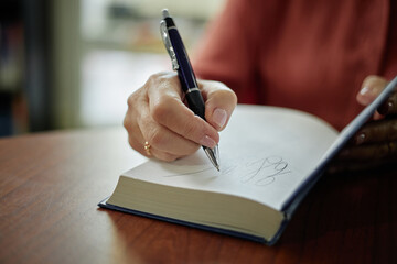 Hands of female author signing book with pen, close up of writing autograph on blank page, wooden...