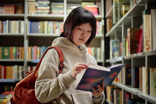 Asian teenager girl standing in library reading book, wearing backpack, focusing on pages, surrounded by shelves filled with books, engaging in independent study - Powered by Adobe