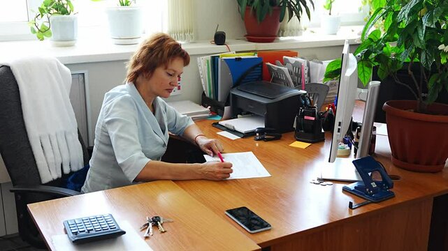 Woman working at a cluttered office desk with folders, documents, and plants in the background. Concept of everyday workflow, paperwork routine, and real administrative work environment