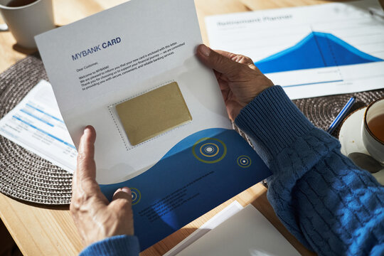 Senior person holding financial document featuring bank card sleeve over wooden table with graphs and papers. Gesture indicating careful analysis of financial data - Powered by Adobe