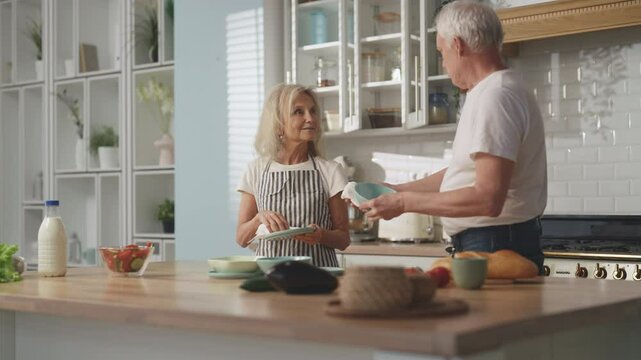 Do housework together, elderly spouses wiping dishes after washing and chatting. Happy married couple in old age, calm and joyful aged people spending weekend at home, wash kitchenware, serving table