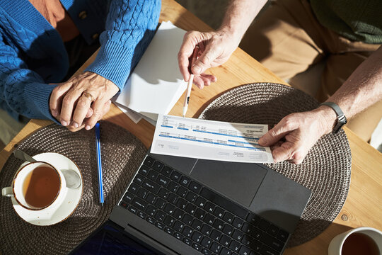 Hands of senior husband and wife studying household bill, while reviewing monthly utility expenses and financials at kitchen table, with laptop and tea cup - Powered by Adobe