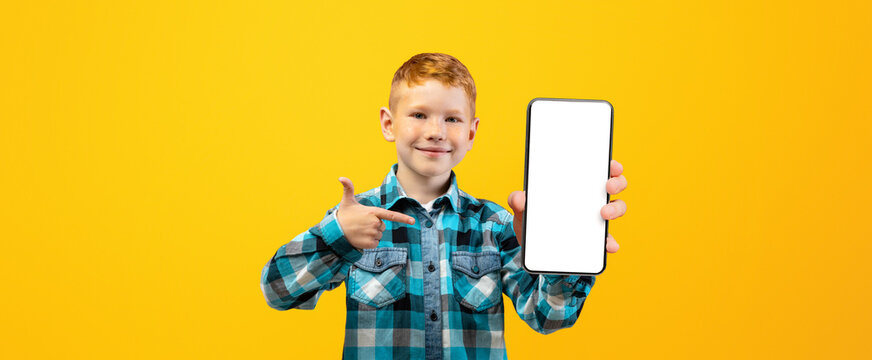 Smiling ginger boy in casual shirt pointing at modern mobile phone with empty screen over yellow studio background, mockup. Happy schoolboy with smartphone for education or entertainment, copy space