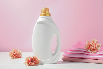 Bottle of fabric softener, clean clothes and carnation flowers on white wooden table against pink background