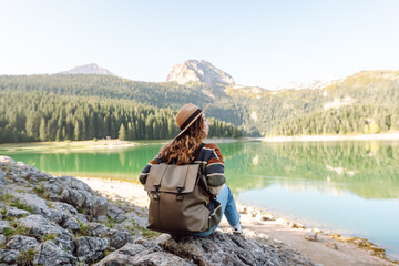 Rear view of female traveler wearing hat and carrying backpack enjoying mountain lake. Young woman...