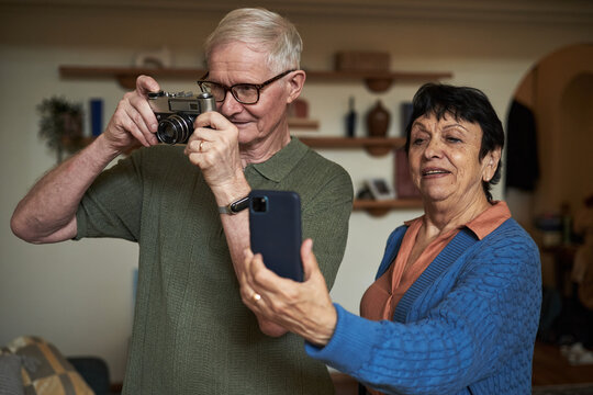 Elderly couple engaging in photography, using a vintage camera and a smartphone for capturing moments together in a cozy living room setting
