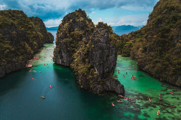 Tourists Kayaking Through the Turquoise Waters of Big Lagoon with impresive cliffs and bangka boats moored in a sunny day in Minoloc Island, El Nido, Palawan, Philippines