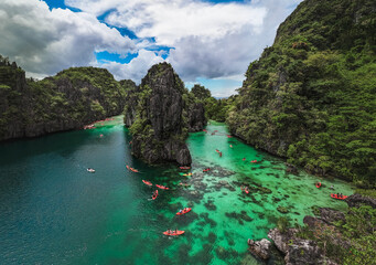 Tourists Kayaking Through the Turquoise Waters of Big Lagoon with impresive cliffs and bangka boats moored in a sunny day in Minoloc Island, El Nido, Palawan, Philippines