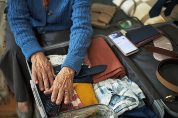 Senior person packing luggage with various essentials including clothing, gadgets, and documents, preparing for travel on vacation. Focus on hands organizing items in suitcase with care