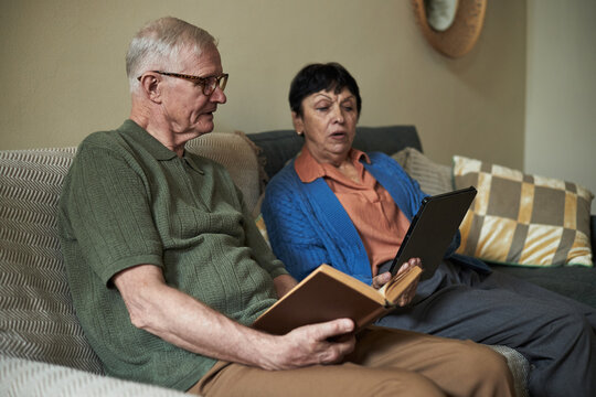 Senior couple sitting on sofa, engaged in reading with one using tablet and other holding book in cozy living room. Mirror and patterned pillows adorn background