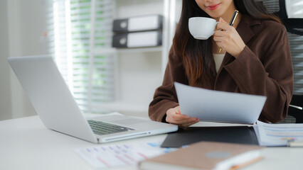 Close-up of an Asian businesswoman holding a cup of coffee and reviewing documents at her desk with a laptop in a modern office setting.