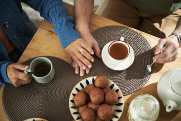 Senior couple's hands showing gentle touch while enjoying tea around rustic wooden table. Tea cups, saucers, and biscuits with warm, inviting atmosphere