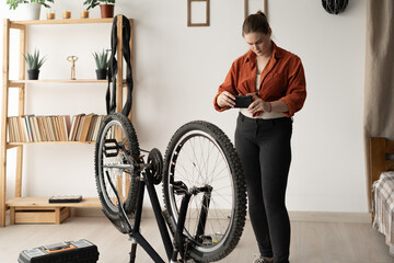 Young woman standing next to her bicycle at home taking a photo of it with her smartphone. DIY...