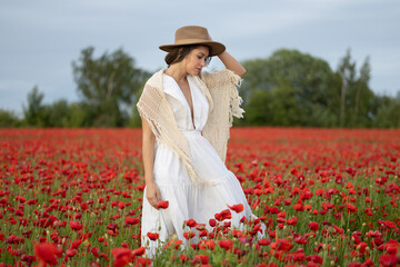 Young woman in a white dress and hat walks among summer poppies