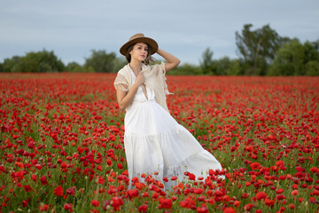 Young woman in a white dress and hat posing among summer poppies