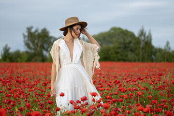 Young woman in a white dress and hat posing among summer poppies