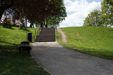 serene park path leads to sunlit stone steps, flanked by lush green grass and trees under a partly...