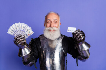 Smiling mature man in medieval knight's armor holding dollar bills and credit card against purple...