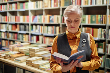 Caucasian senior woman reading book while standing in front of bookshelves filled with books in...
