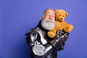 Elderly man wearing shiny medieval armor holding a teddy bear on a purple background, highlighting...