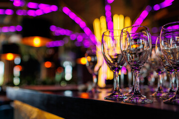 Empty clear wineglasses stand in rows on bar counter at nightclub, blurred purple neon and yellow lights on background. Clean cocktail glasses ready for celebrate party.