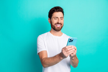 Happy young man using smartphone in casual white shirt with teal background, smiling while holding...