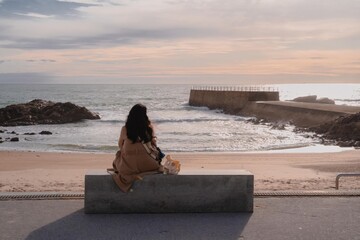 Woman in coat sitting on a bench, gazing at the ocean and harbor wall under soft sunset sky