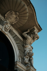 Stone figure holding a cornice on an ornate building in Vrsac, Serbia, with symbolic facial features and scroll design.