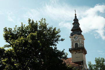Baroque style clock tower of a church in Vrsac, Serbia country, surrounded by green tree branches against a blue sky.