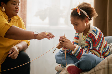 Young Black girl learning knitting while spending quality time with grandmother in living room,...