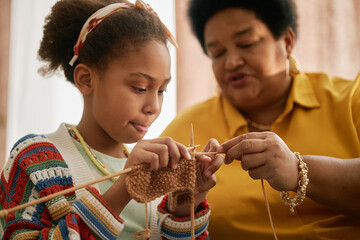 Focused African American young girl learning knitting while grandmother teaching child craft technique at home
