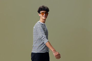 Smiling man wearing striped shirt and sunglasses stands against olive background expressing casual...