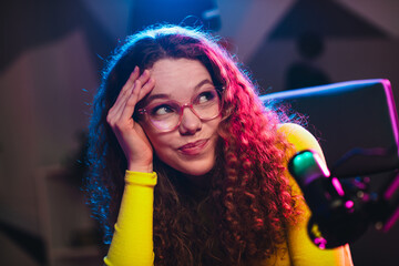 Young woman with curly hair in neon lights looking thoughtful while streaming, surrounded by...
